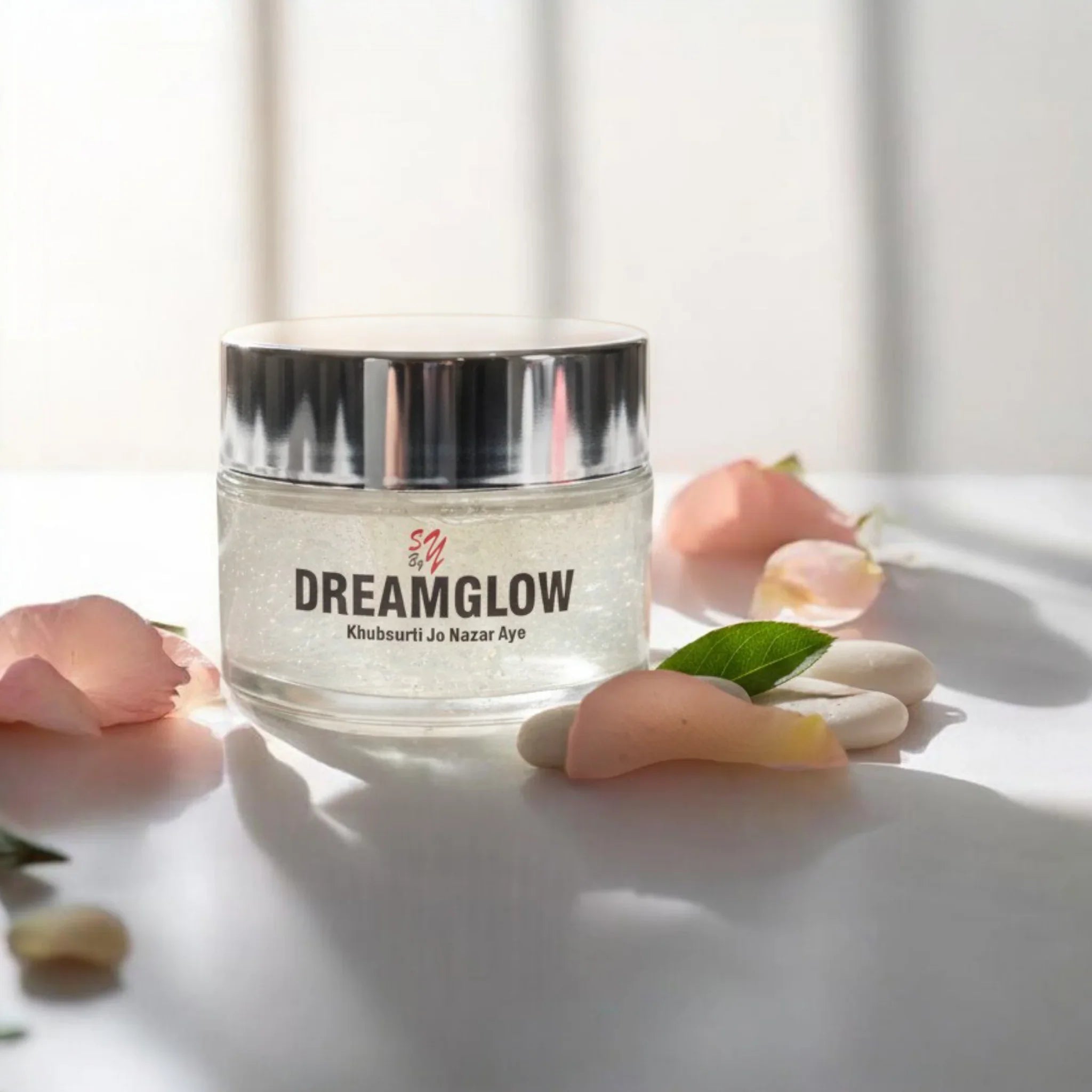 Glass jar of DreamGlow face cream on white surface with rose petals and leaves